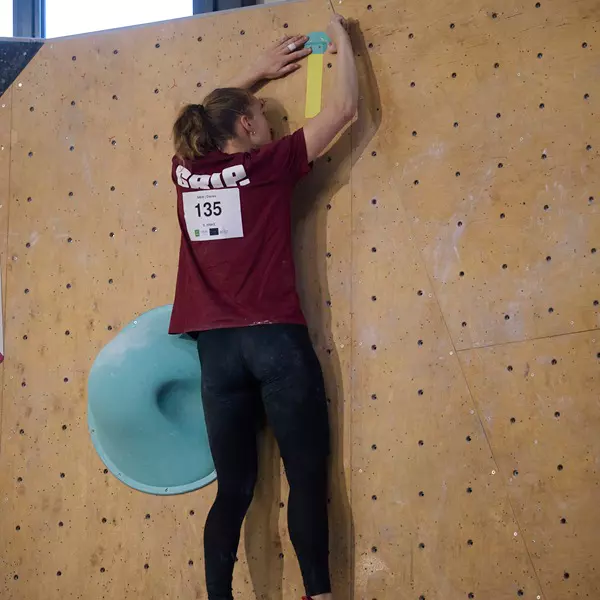 Iemand in een rood shirt met het nummer 135 beklimt een indoor-bouldermuur en reikt met beide handen naar een gele greep. Vlak bij zijn of haar benen bevindt zich een grote blauwe greep.